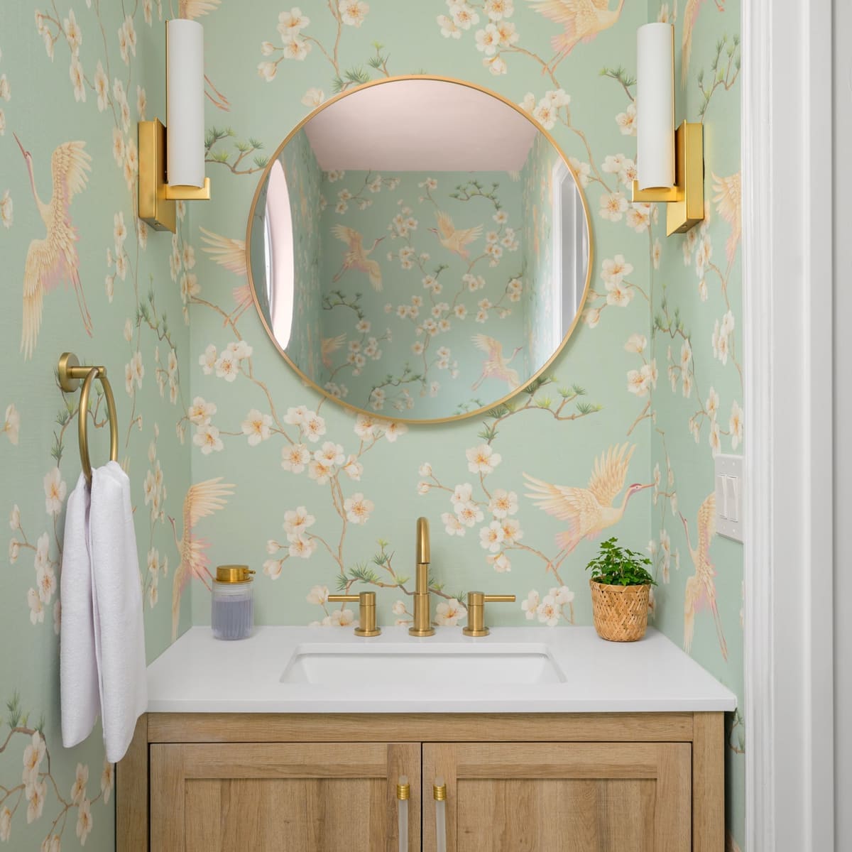 Powder room with mint crane and blossom wallpaper, a round brass mirror, brass sconces and light oak vanity