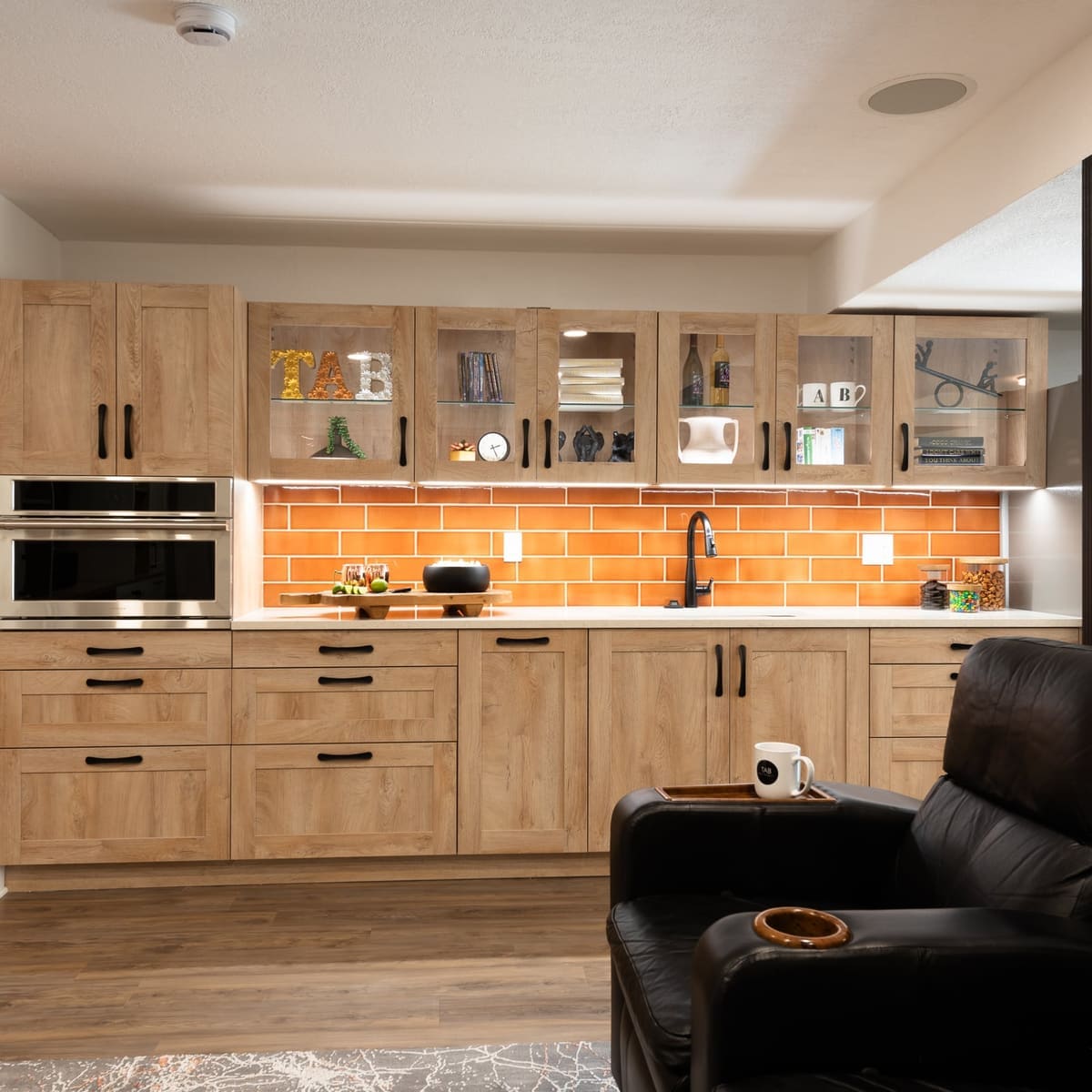 Basement kitchenette with light oak shaker cabinets, glass uppers, orange glazed subway tile and black hardware